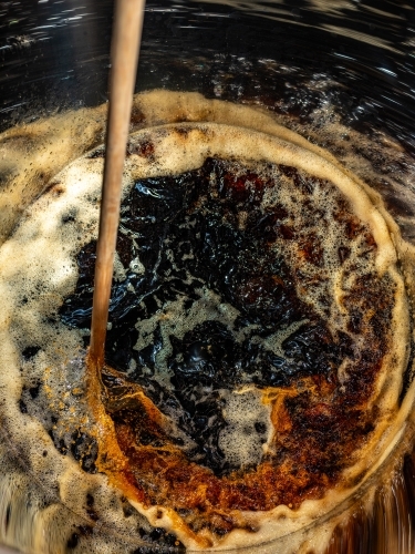 A dark liquid with frothy bubbles and swirls stirred with a wooden paddle - Australian Stock Image