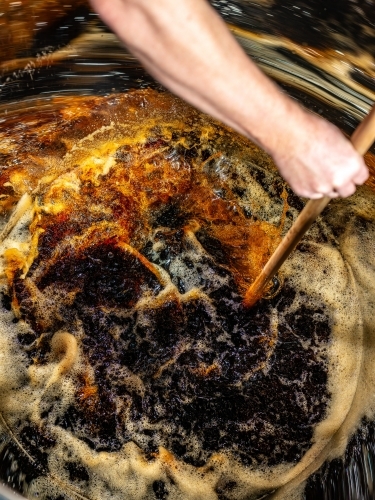 A dark liquid with frothy bubbles and swirls stirred with a wooden paddle - Australian Stock Image