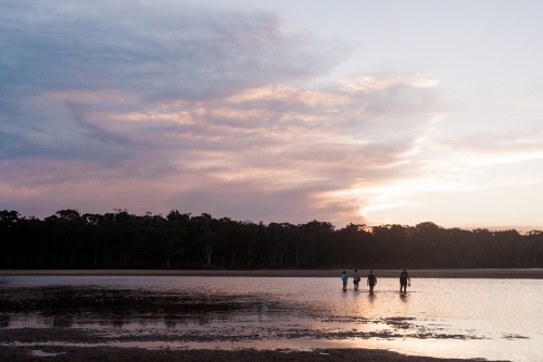 A family of four walk through a swallow lake with fishing gear at dusk.