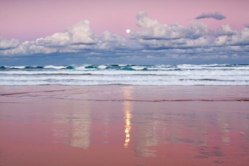 A full Moon rising out of clouds over a beach with colourful reflections on the wet sand