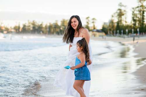 A girl and her mother laugh and splash in the waves at the beach in Australia on a sunny day. - Australian Stock Image