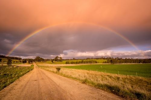 A ground level view looking down a dirt road with a vivid rainbow arching overhead