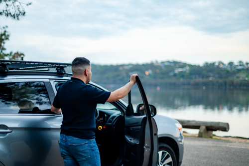 A happy man in his 30s stands by his car near a peaceful lake in a park, looking away - Australian Stock Image
