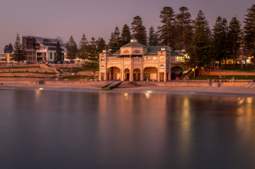 A heritage beachside pavilion glowing at dusk reflected in calm water framed by Norfolk pines - Australian Stock Image