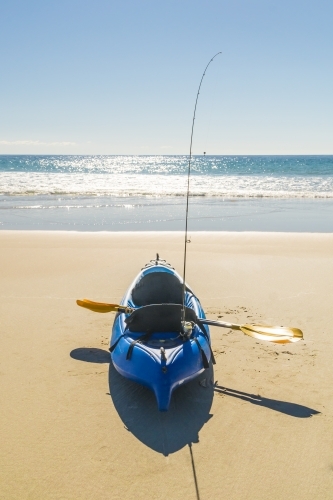 A kayak sitting on a beach with a paddle and fishing rod on board