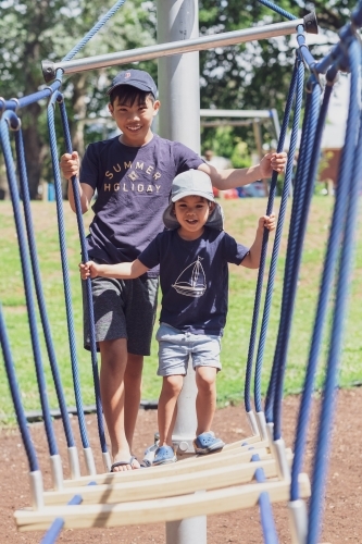 a kid with his older brother crossing a wobbly bridge at the playground - Australian Stock Image