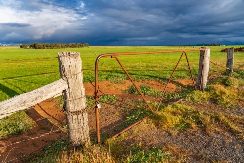 A locked farm gate and fence posts with green farmland under dark clouds