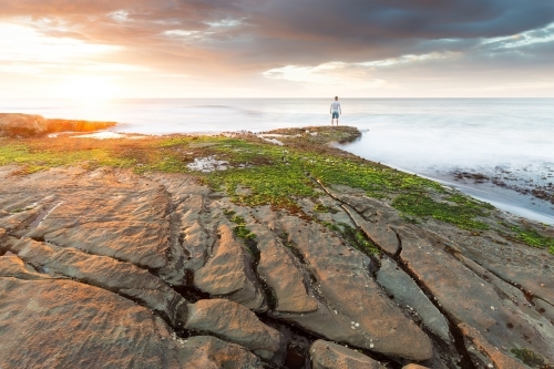 A lone figure standing on a rock ledge looking out at the ocean at sunrise