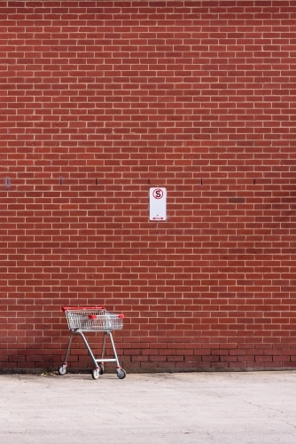 A lonely shopping trolley stays in a non-stopping open space, against a red brick wall.