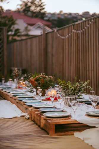 A long wooden table adorned with elegant tableware and glassware is set for an outdoor dinner - Australian Stock Image