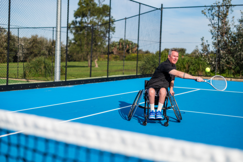 A male athlete using a wheelchair swings a tennis racquet while playing on a blue court - Australian Stock Image