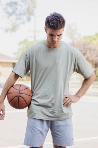 A male in his twenties holding a basketball / taking a break on an outdoor basketball court