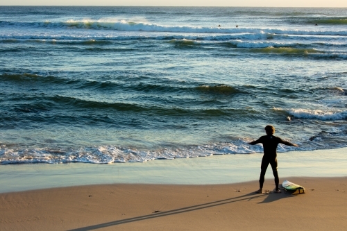 A male surfer in a wetsuit warms up with his surfboard beside him on the beach at Cabarita. - Australian Stock Image