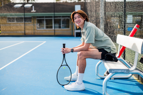 A male tennis player relaxes on the bench, holding a racquet after training on a bright day. - Australian Stock Image