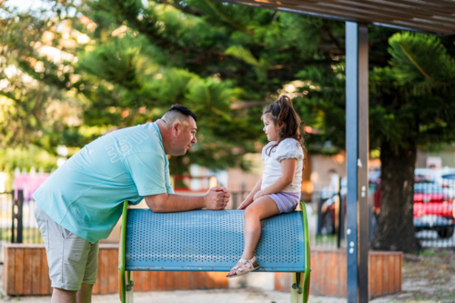 A man and a young girl talk at a playground near trees in Australia at sunset. - Australian Stock Image