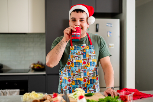 A man in a colourful apron enjoys a drink while cooking a festive lunch in his kitchen - Australian Stock Image