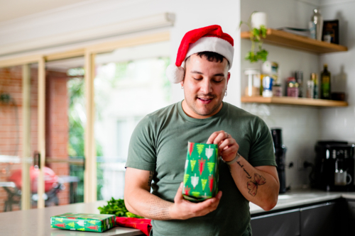 A man in a Santa hat excitedly unwraps a colourful present at home, celebrating Christmas - Australian Stock Image