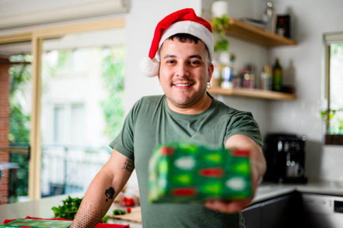 A man in a Santa hat smiles as he holds out a wrapped gift at his home for Christmas festivities - Australian Stock Image