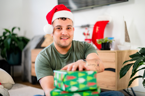 A man in a Santa hat smiles while holding Christmas gifts in a cosy loungeroom - Australian Stock Image