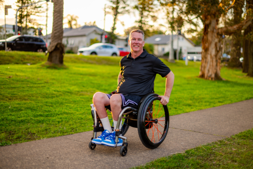 A man in a wheelchair is smiling while rolling along a footpath in a park at sunset - Australian Stock Image