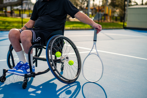 A man in a wheelchair on a tennis court holding a racquet with tennis balls nearby. - Australian Stock Image