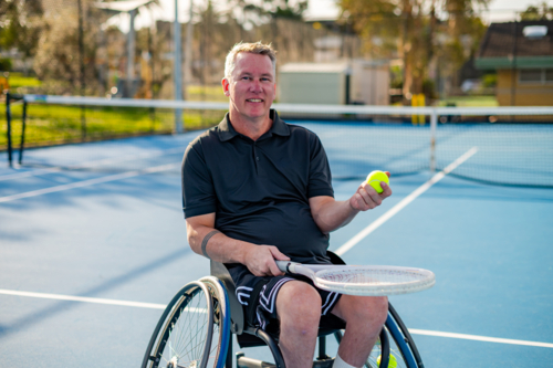 A man in a wheelchair smiles while holding a tennis ball on an outdoor blue court - Australian Stock Image