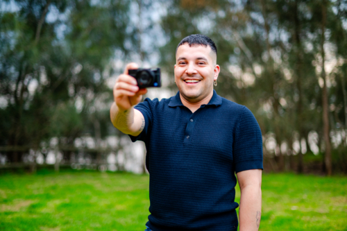 A man in his 30s happily takes a photo with his camera while standing in a grassy park. - Australian Stock Image