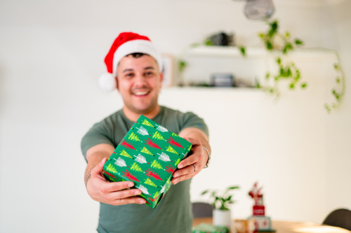 A man in his 30s in a Santa hat joyfully presents a gift while celebrating Christmas. - Australian Stock Image