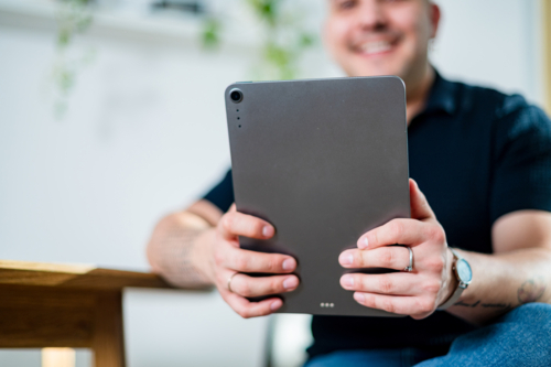 A man in his 30s smiles while holding a tablet at a wooden table in a cosy home setting - Australian Stock Image