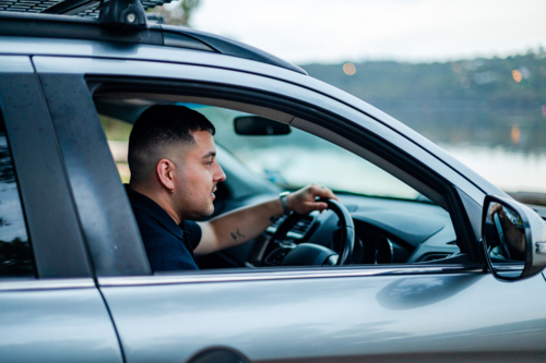 A man sits in a car, focused on the road beside a tranquil lake in a park during daylight. - Australian Stock Image