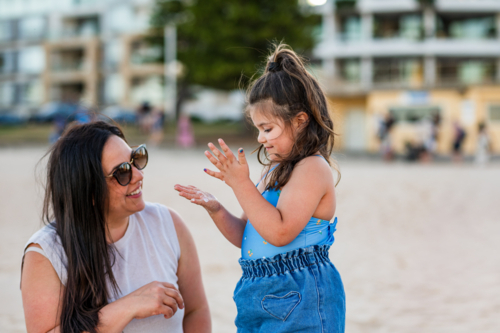 A mother smiles at her young daughter playing with sand at the beach. - Australian Stock Image