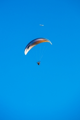A paraglider floats in the clear blue sky with a distant aircraft above and in the background. - Australian Stock Image