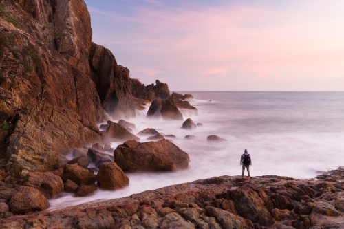 A person is silhouetted by a pink sunrise at the base of a coastal cliff in a beautiful seascape.