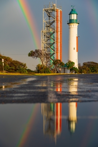 A rainbow arching over a lighthouse and signal towers with reflections in foreground puddles - Australian Stock Image