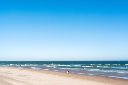 A senior couple walks along a wide, long beach on summers day with a big blue sky overhead.