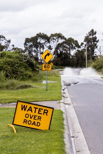 A sign which says 'Water over road' warning of flash flooding, there is a car driving through it - Australian Stock Image