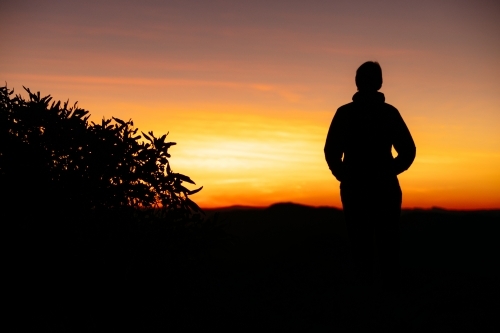 A silhouetted female stands with a vibrant coloured sunrise in front of her. - Australian Stock Image