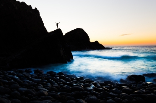 A silhouetted person welcomes the sunrise on a beach of boulders in a beautiful coastline seascape. - Australian Stock Image