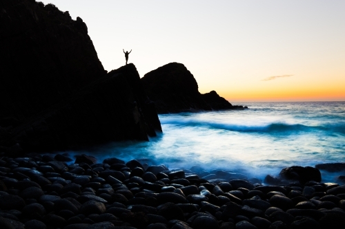 A silhouetted person welcomes the sunrise on a beach of boulders in a beautiful coastline seascape.