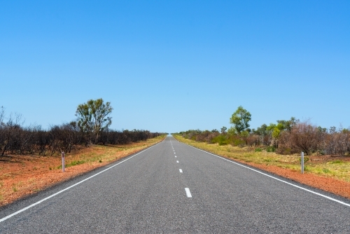 A straight bitumen road through low vegetation and orange soil with dotted white line and blue sky - Australian Stock Image