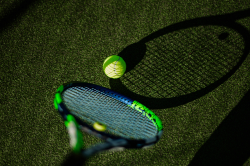 A tennis racquet hovers close to a vibrant yellow ball, both resting on lush green turf - Australian Stock Image