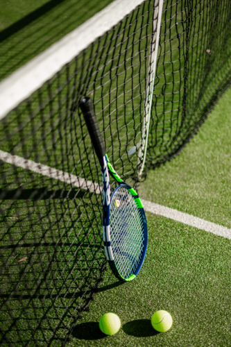 A tennis racquet leans against a net with two tennis balls on the green court surface - Australian Stock Image