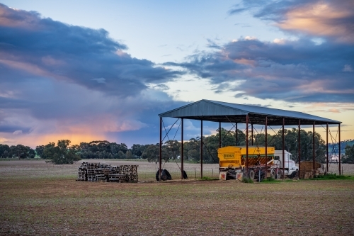 A truck and farm machinery under a hay shed on farmland at sunset - Australian Stock Image
