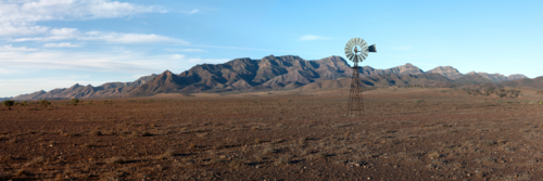 A vintage windmill stands on a stony plain against a rugged mountain backdrop - Australian Stock Image