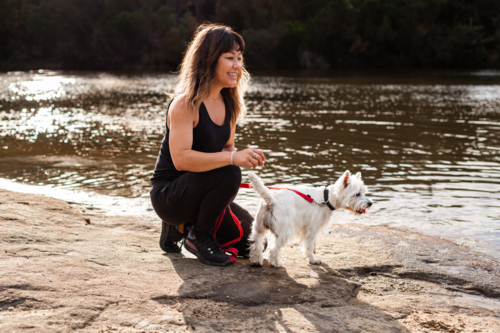 A woman crouches beside a riverbank, smiling as she interacts with her little dog near the water. - Australian Stock Image