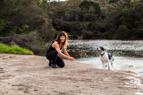 A woman enjoys a playful moment with her dog at a calm riverbank enjoying nature. - Australian Stock Image