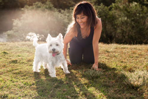 A woman enjoys time outdoors with her small white dog on a sunny day in a park - Australian Stock Image