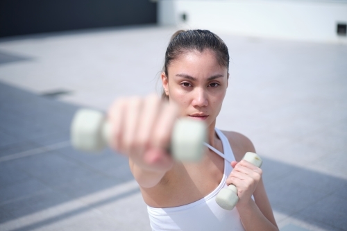 A woman in a white tank top and pink leggings working out using white dumbbells - Australian Stock Image
