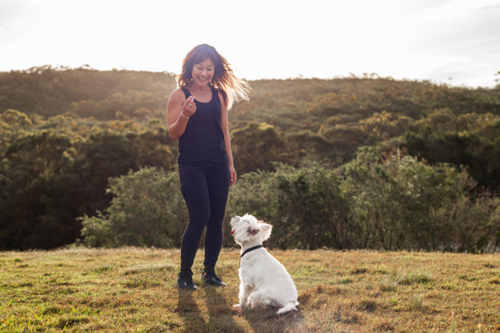 A woman in activewear trains her dog at a park during sunset, surrounded by nature - Australian Stock Image