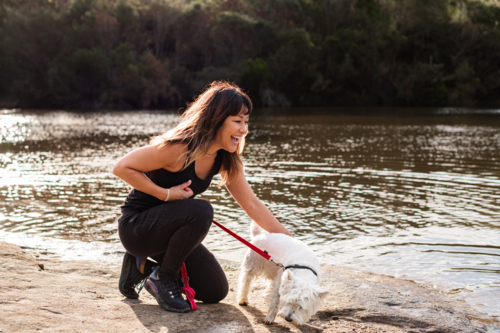 A woman kneels by the riverbank, laughing as she plays with her dog on a sunny afternoon - Australian Stock Image